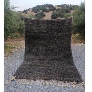 Puede incluir: Una alfombra tejida a mano de color gris oscuro con un borde de flecos. La alfombra se muestra al aire libre sobre una superficie de grava.