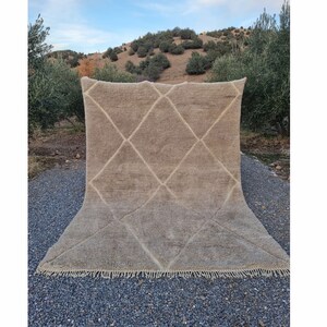 May include: A large, off-white, handwoven rug with a diamond pattern. The rug has a fringe border and is laid out on a gravel surface.