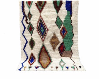May include: A white Moroccan rug with a geometric pattern in shades of red, blue, green, and brown. The rug has a diamond-shaped design with a fringe border.