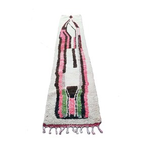 May include: A long, white rug with a geometric design in pink, green, and brown. The rug has fringe on the ends.