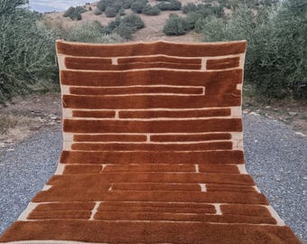 Op de afbeelding: Een groot, rechthoekig tapijt met een bruin, geometrisch patroon van horizontale en verticale lijnen. Het tapijt heeft een crèmekleurige achtergrond en een gefranjerde rand. Het tapijt wordt buiten op een grindoppervlak getoond, met bomen en een heuvel op de achtergrond.