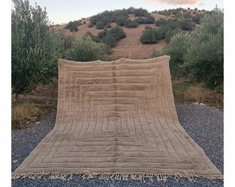Puede incluir: Una alfombra grande, rectangular, de color beige con un patrón geométrico. La alfombra tiene un borde de flecos y está colocada sobre una superficie de grava.