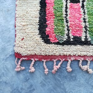 May include: A close-up of a colorful Moroccan rug with a fringe border. The rug has a geometric pattern with pink, green, black, and white stripes.
