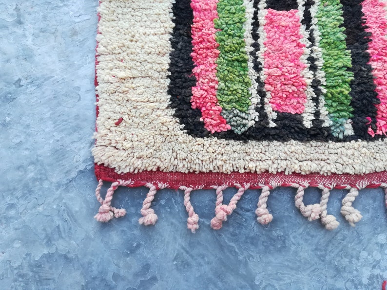 May include: A close-up of a colorful Moroccan rug with a fringe border. The rug has a geometric pattern with pink, green, black, and white stripes.