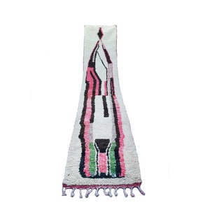 May include: A long, white rug with a geometric design in pink, black, and green. The rug has a fringe on the bottom.