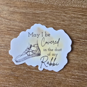 May include: A white sticker with a yellow watercolor background and black text that reads "May I be covered in the dust of my Rabbi". A black and white drawing of a sandal is on the sticker.