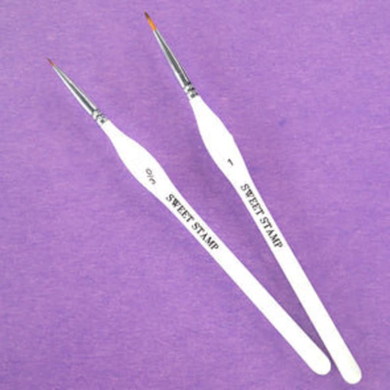 May include: Two white paint brushes with black lettering that reads "Sweet Stamp" and the size of the brush. The brushes have a fine point.
