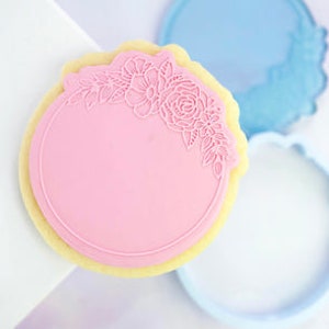 Puede incluir: Un cortador de galletas rosa con un diseño floral alrededor del borde. El cortador tiene forma de círculo con un centro en blanco.