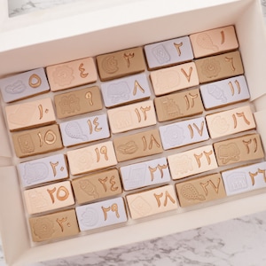Peut inclure: Une boîte de 36 petits biscuits rectangulaires décorés de symboles islamiques et de chiffres en or et blanc. Les biscuits sont disposés en rangées et se trouvent dans une boîte blanche sur un fond brun clair.