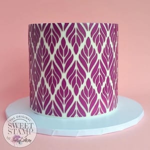 May include: A white cake with a purple leaf pattern design. The cake is on a white cake board. The text on the cake reads "The Original Sweet Stamp by Amy Cakes". The background is a solid pink colour.