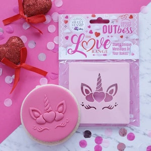 Puede incluir: Una galleta rosa con un diseño de unicornio estampado en ella. El unicornio tiene un cuerno, orejas y tres corazones para una melena. La galleta está al lado de una plantilla de plástico con el mismo diseño de unicornio. La plantilla está empaquetada en un envoltorio blanco con el texto "The Love Range" y "Sweet Stamp Outboss" impreso en ella.