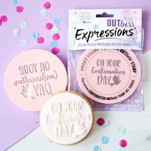 Puede incluir: Dos cortadores de galletas rosas y blancos con el texto "On Your Confirmation Day" y un diseño de paloma. Los cortadores de galletas están empaquetados en una bolsa de plástico transparente con el texto "OUTboss Expressions" y "Sweet Stamp" en el empaque.
