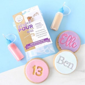 Puede incluir: Un conjunto de tres galletas decoradas con los nombres "Elle", "Ben" y el número "13" en glaseado dorado. Las galletas están al lado de una bolsa de plástico transparente que contiene un juego de plantillas de plástico transparente. Hay dos botellas de glaseado de colores, una rosa y una dorada, con una punta de aguja.