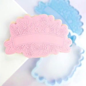 Op de afbeelding: Roze plastic koekjesvormpje met een bloemmotief en een lege banner in het midden. De vormpje is ontworpen om koekjes te maken met een bloemrand en een banner om tekst toe te voegen.