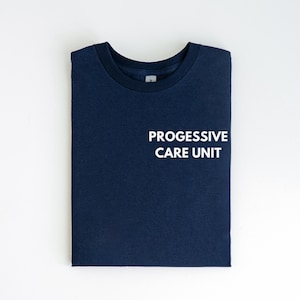 May include: A folded navy blue t-shirt with the white text "PROGRESSIVE CARE UNIT" on the front. The shirt is made of a soft material and has a crew neck.