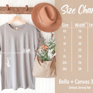 May include: Size chart for a unisex jersey tee, showing measurements in inches for width and length. The chart includes sizes XS through 3XL. The image also shows a gray t-shirt hanging on a wooden hanger, a brown hat, and a brown bag.