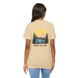 May include: A tan t-shirt with a graphic of a lake scene with a campfire and two chairs. The graphic also includes a yellow sun, green trees, and a pink mountain. The text "LEGEND LAKE 2024" is printed below the graphic.