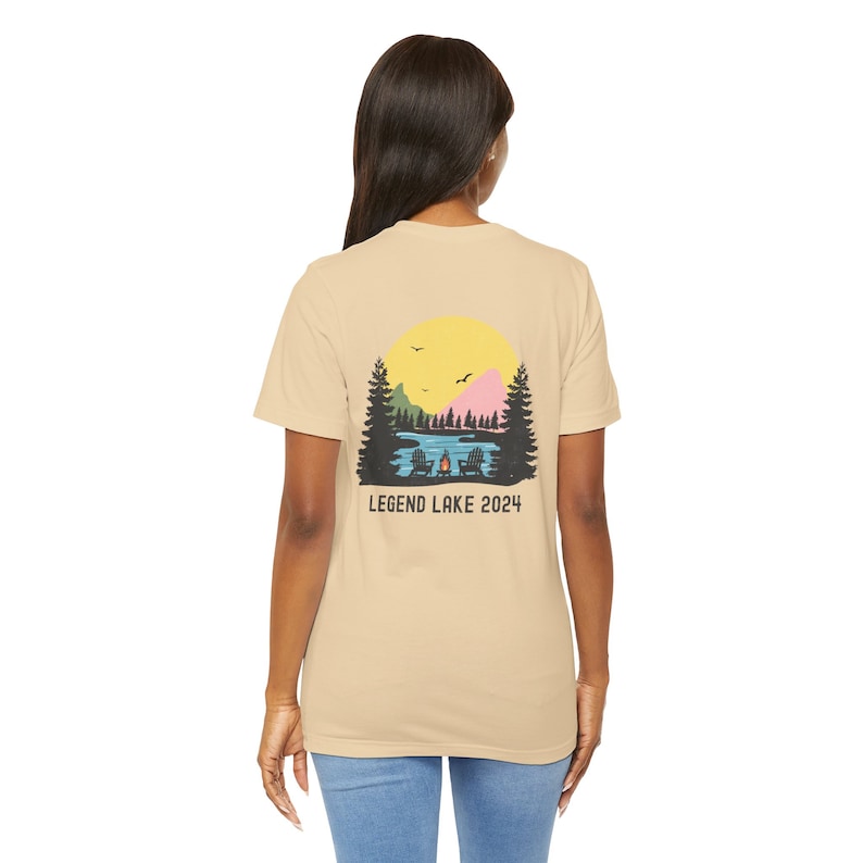 May include: A tan t-shirt with a graphic of a lake scene with a campfire and two chairs. The graphic also includes a yellow sun, green trees, and a pink mountain. The text "LEGEND LAKE 2024" is printed below the graphic.