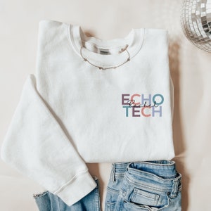 Puede incluir: Una sudadera blanca de cuello redondo con el texto "ECHO Rachel TECH" en un diseño colorido apilado. Un delicado collar dorado descansa sobre el cuello. La sudadera se combina con unos vaqueros azules.