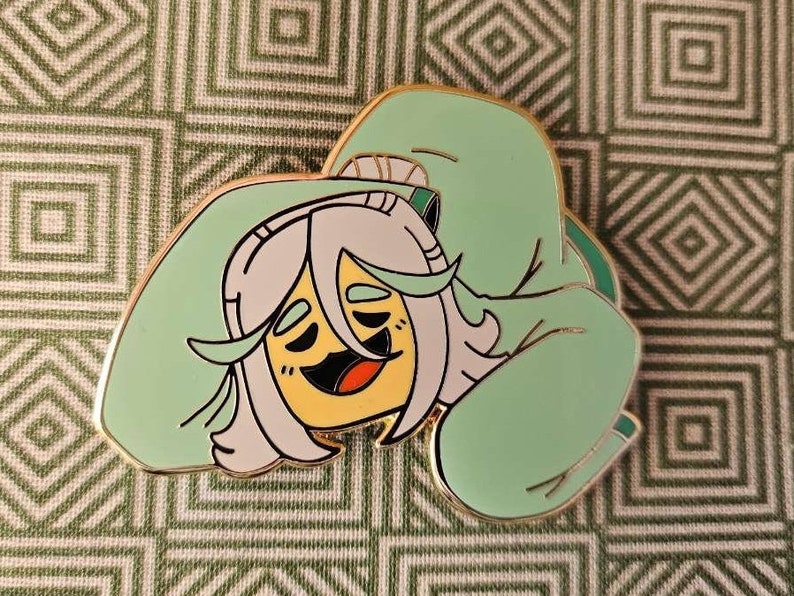 Peut inclure: Une &eacute;pingle en &eacute;mail verte repr&eacute;sentant un personnage de dessin anim&eacute; aux cheveux blancs et au visage jaune, portant un sweat &agrave; capuche vert. Le personnage sourit et a les yeux ferm&eacute;s, comme s'il dormait.