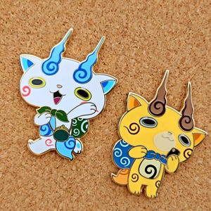 以下が含まれることがあります： 2つのエナメルピン。漫画の猫のようなキャラクターが描かれています。1つは青いアクセントの白、もう1つは茶色のアクセントの黄色です。どちらも角とバッグがあり、笑顔です。ピンは金色の縁取りです。