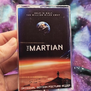 May include: A clear plastic cassette case featuring the soundtrack for The Martian. The cover art displays a view of Earth from space above a red Martian landscape with a lone figure standing on a ridge. Text reads: Help is only 140 million miles away.