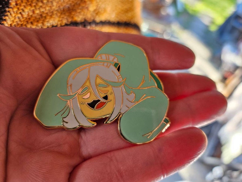 Peut inclure: Une &eacute;pingle en &eacute;mail verte et jaune repr&eacute;sentant un personnage de dessin anim&eacute; avec des cheveux blancs et un sweat-shirt vert. Le personnage sourit et a un visage jaune.