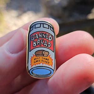 May include: A gold-colored enamel pin in the shape of a can of bread. The pin features the words "CANNED BREAD" in red and white, with an illustration of bread and the text "BEST THING SINCE SLICED".