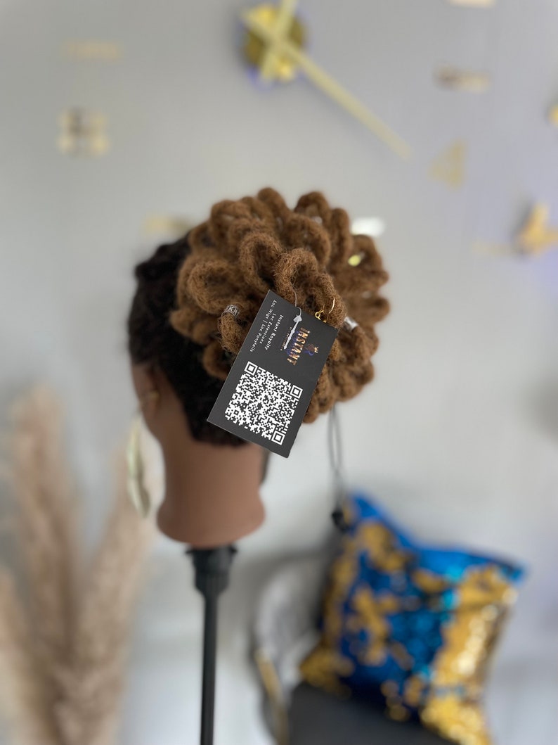 May include: A brown wig styled in a bun, displayed on a mannequin head. The wig has a textured appearance and is adorned with small, silver-colored accents. A tag with a QR code and the word "INSTANT" is attached.