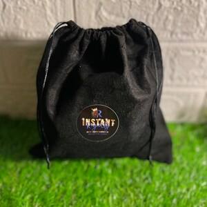 May include: A black drawstring bag with a round sticker that reads "INSTANT ROYALTY LOC EXTENSIONS" in gold and white. The bag is made of a soft, dark material and sits on a bed of green grass.
