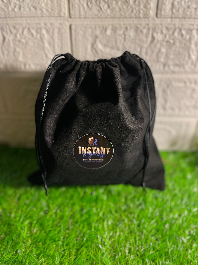 May include: A black drawstring bag with a round sticker that reads "INSTANT ROYALTY LOC EXTENSIONS" in gold and white. The bag is made of a soft, dark material and sits on a bed of green grass.