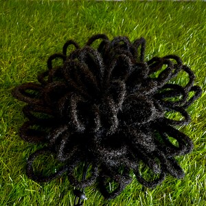 May include: A black hair accessory, likely a hair wrap or bun, is displayed against a backdrop of green grass. The accessory is made of numerous looped strands, creating a textured, voluminous appearance. The accessory is designed to be worn in the hair.
