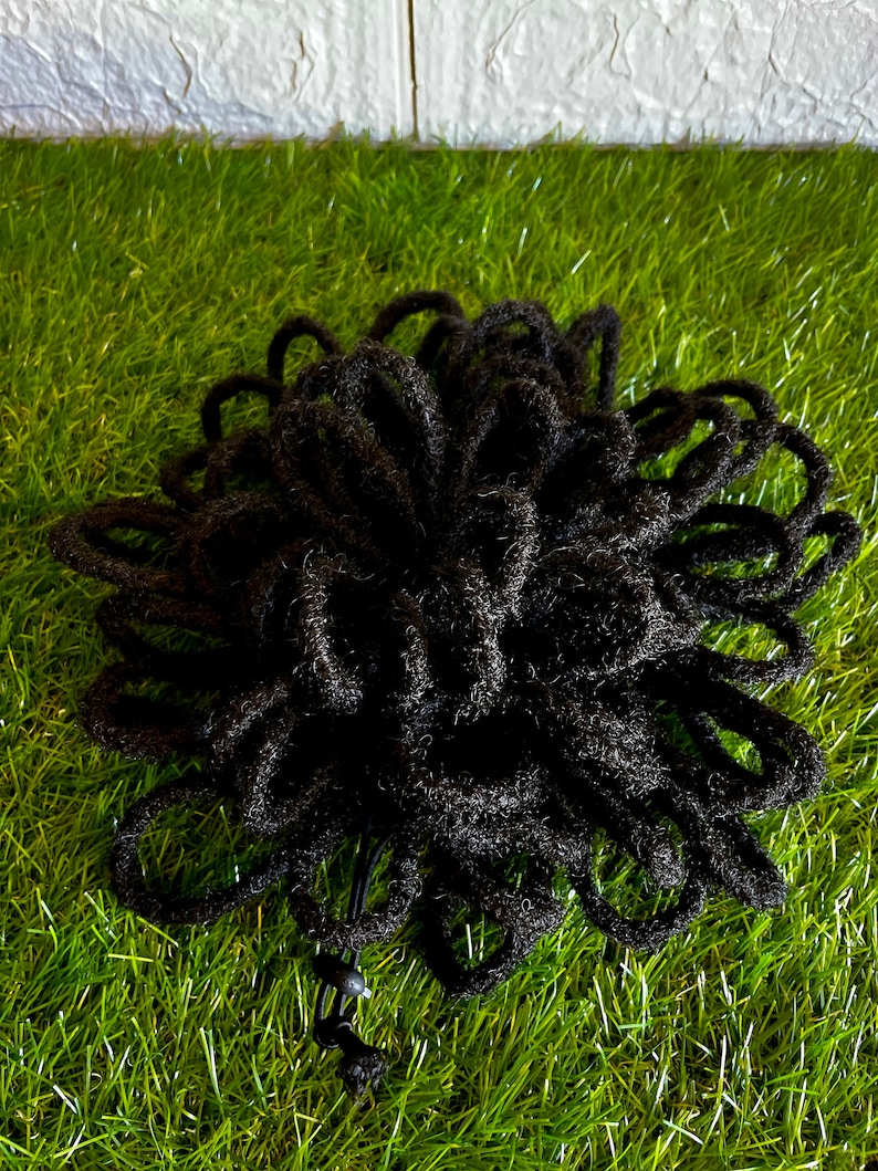 May include: A black hair accessory, likely a hair wrap or bun, is displayed against a backdrop of green grass. The accessory is made of numerous looped strands, creating a textured, voluminous appearance. The accessory is designed to be worn in the hair.