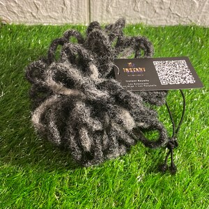 May include: A bundle of gray and white dreadlock extensions. A black tag attached reads "Instant Royalty" with a QR code. The extensions are on a bed of green artificial grass.