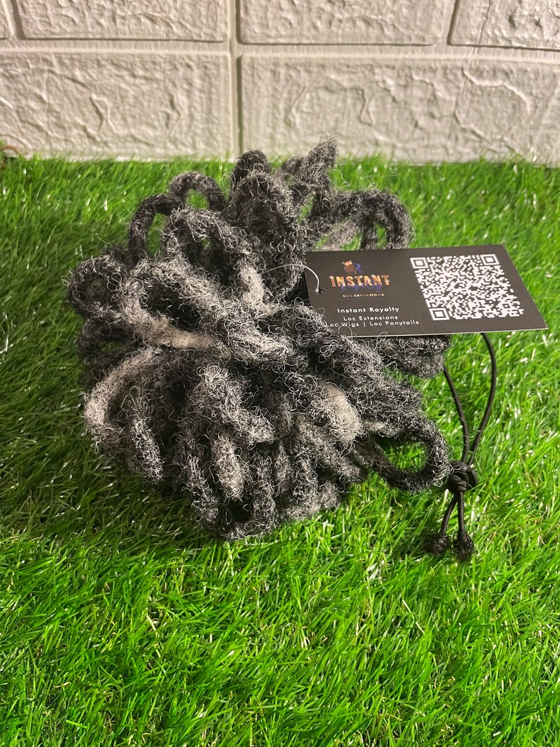 May include: A bundle of gray and white dreadlock extensions. A black tag attached reads "Instant Royalty" with a QR code. The extensions are on a bed of green artificial grass.