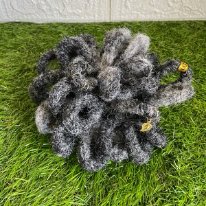 May include: A bundle of gray and black dreadlocks with gold-colored accents. The dreadlocks are arranged in a circular shape and are made of a textured material. A small gold leaf charm is attached to one of the dreadlocks.