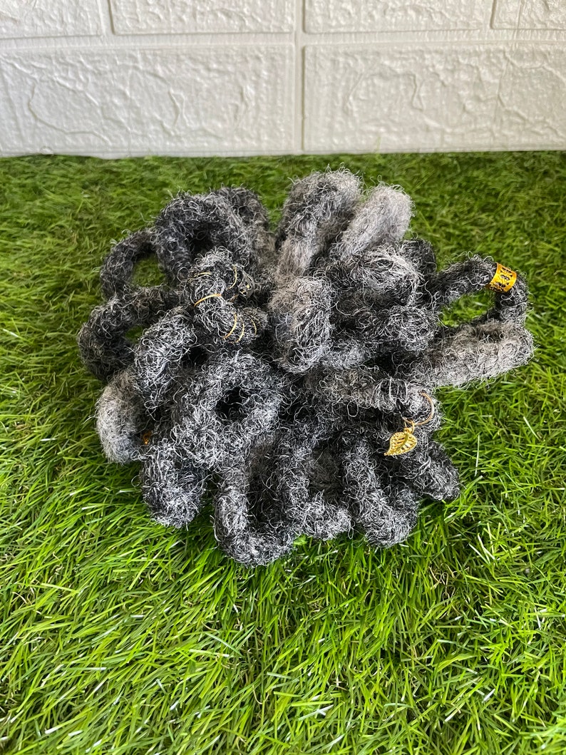 May include: A bundle of gray and black dreadlocks with gold-colored accents. The dreadlocks are arranged in a circular shape and are made of a textured material. A small gold leaf charm is attached to one of the dreadlocks.