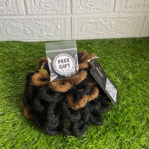 May include: A bundle of black and brown dreadlocks, with a small gold-colored charm and a clear bag labeled "FREE GIFT for you." The dreadlocks are arranged on a bed of green grass.