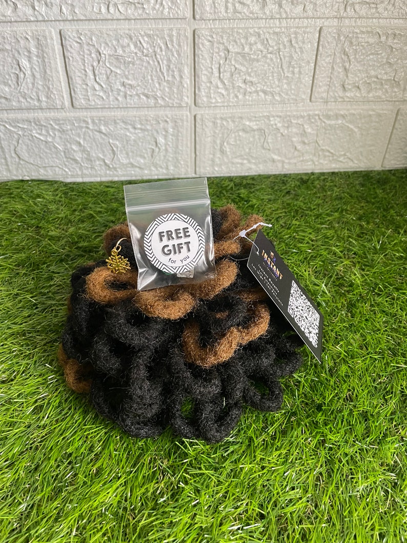 May include: A bundle of black and brown dreadlocks, with a small gold-colored charm and a clear bag labeled "FREE GIFT for you." The dreadlocks are arranged on a bed of green grass.