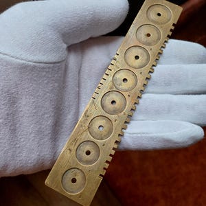May include: A vintage brass gauge with multiple circular holes of varying sizes. The gauge has etched markings and numbers along its edge. The tool is held by a gloved hand, suggesting careful handling and preservation of the antique item.