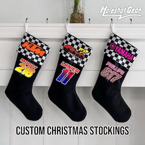May include: Three black Christmas stockings with white hanging loops. Each features a checkered cuff and personalized graphics: KTM with Moody 216, Cobra Moto with Lanier 11, and Yamaha with Phillips 617. The text Holeshot Gear appears in the top right corner.