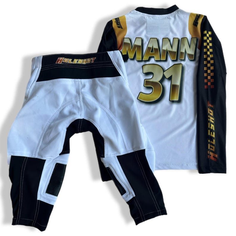 May include: White and black motocross gear set with a long-sleeved jersey and pants. The jersey has the name "MANN" and the number "31" printed on the back in gold. The jersey and pants both have the word "HOLESHOT" printed on the side.