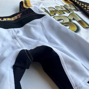 May include: White and black athletic shorts with a black waistband and the word "LESHOT" printed on the waistband. The shorts have a black mesh panel on the side and a white jersey with the word "IANN" and the number "31" printed on it is visible in the background.