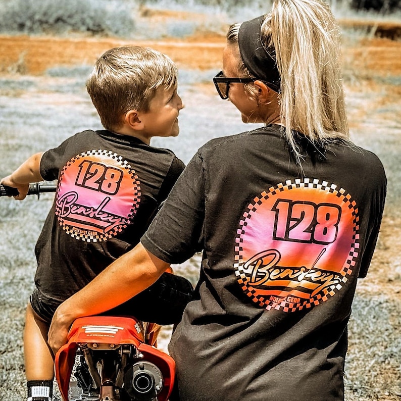 Puede incluir: Dos personas con camisetas negras con un gr&aacute;fico circular de color rosa y naranja. El gr&aacute;fico presenta el n&uacute;mero 128 y la palabra "Berkeley" en una fuente blanca de escritura. El c&iacute;rculo est&aacute; rodeado por un patr&oacute;n a cuadros.