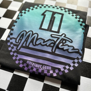Puede incluir: Camiseta negra con un dise&ntilde;o circular que presenta un degradado de azul a morado, el n&uacute;mero "11" y el texto "Martin" en una fuente retro. El dise&ntilde;o est&aacute; rodeado por un borde a cuadros y el texto "HOLESHOT GEAR" est&aacute; en la parte inferior.