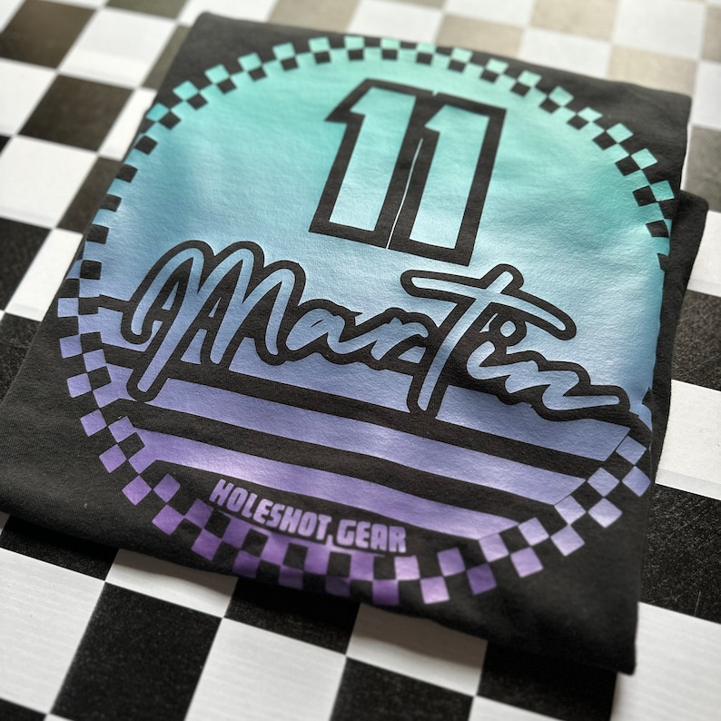 Puede incluir: Camiseta negra con un dise&ntilde;o circular que presenta un degradado de azul a morado, el n&uacute;mero "11" y el texto "Martin" en una fuente retro. El dise&ntilde;o est&aacute; rodeado por un borde a cuadros y el texto "HOLESHOT GEAR" est&aacute; en la parte inferior.