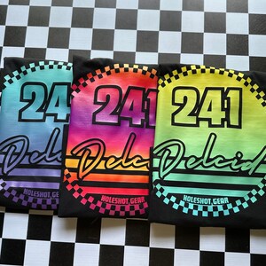 Puede incluir: Tres camisetas negras con un dise&ntilde;o degradado arco&iacute;ris. Cada camiseta presenta un c&iacute;rculo blanco con un contorno negro y el n&uacute;mero "241" en blanco. El texto "Delcid" est&aacute; escrito en una fuente cursiva debajo del c&iacute;rculo. Las camisetas est&aacute;n dispuestas en una fila, con los colores del degradado cambiando de morado a rojo a verde.
