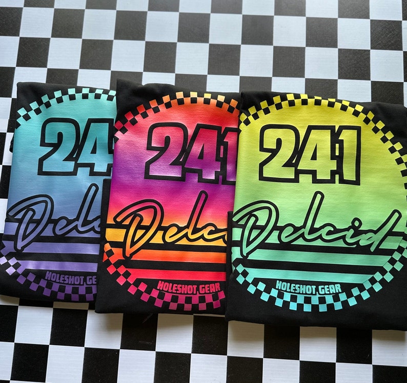 Puede incluir: Tres camisetas negras con un dise&ntilde;o degradado arco&iacute;ris. Cada camiseta presenta un c&iacute;rculo blanco con un contorno negro y el n&uacute;mero "241" en blanco. El texto "Delcid" est&aacute; escrito en una fuente cursiva debajo del c&iacute;rculo. Las camisetas est&aacute;n dispuestas en una fila, con los colores del degradado cambiando de morado a rojo a verde.