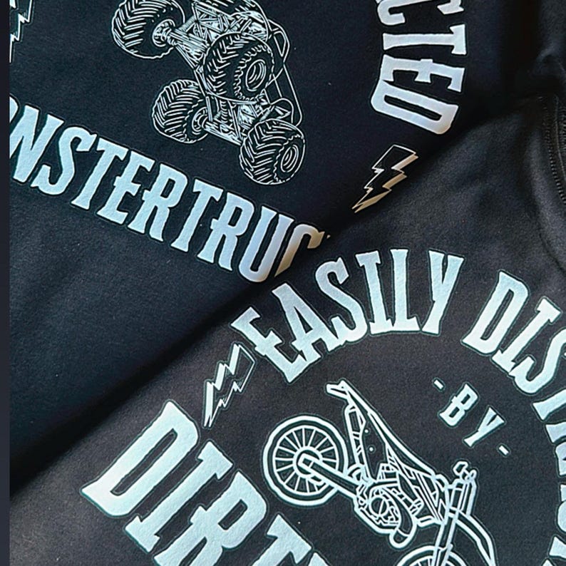 May include: Black t-shirt with white text and graphics. The text reads "Monstertruck Easily Districted - By Dirt". The graphic is a monster truck and a dirt bike.