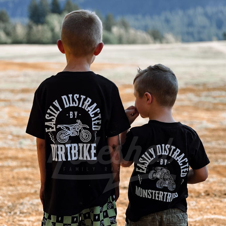 May include: Two children wearing black t-shirts with white graphics. The shirts say "Easily Distracted by Dirtbike" and "Easily Distracted by Monstertruck".